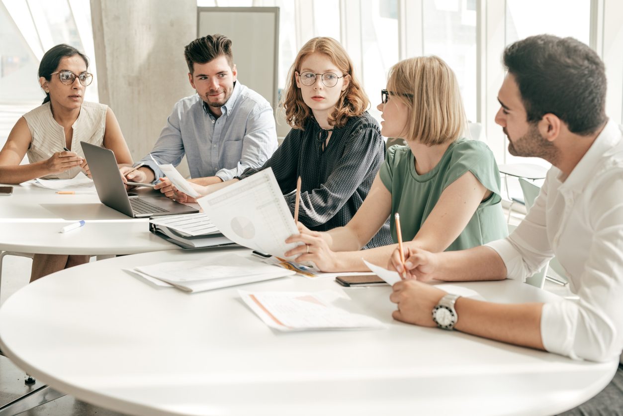A group of five people in a modern office setting discussing documents around a white table, with a laptop and charts in view.