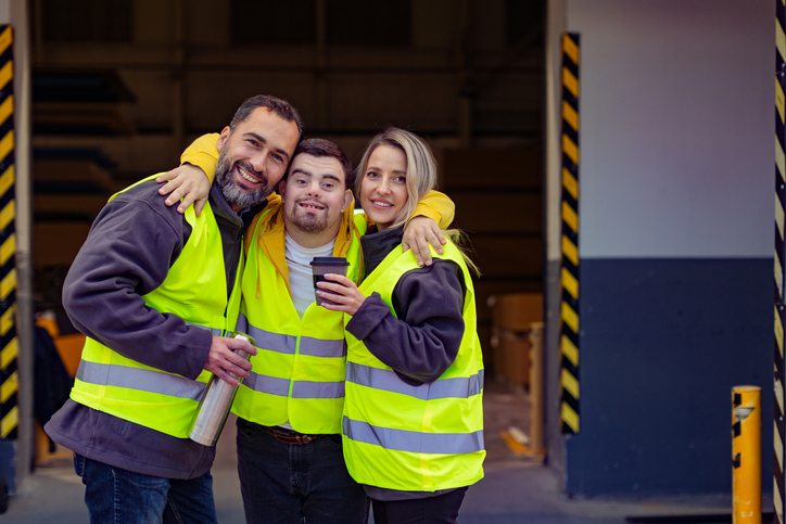 Man with Down syndrome and his colleagues taking break from work, drinking coffee, hot tea from thermos, standing on loading dock. Concept of workers with disabilities, support in workplace.
